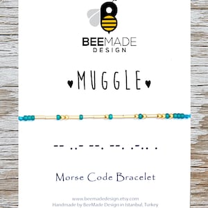 Puede incluir: Una tarjeta blanca con un logotipo de abeja y el texto "BEEMADE DESIGN" y "MUGGLE" en negro. Un brazalete de cuentas turquesa y dorado con la palabra "MUGGLE" en código Morse se muestra en la tarjeta. El texto "Pulsera de código Morse" también está en la tarjeta, junto con la dirección del sitio web y el texto "Hecho a mano por BeeMade Design en Estambul, Turquía".