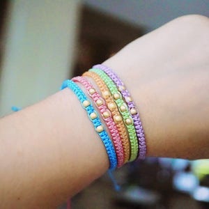 Pu&ograve; includere: Un set di cinque braccialetti dell'amicizia colorati con perline dorate. I braccialetti sono realizzati con cordini sottili e intrecciati in tonalit&agrave; di blu, verde, rosa e viola.