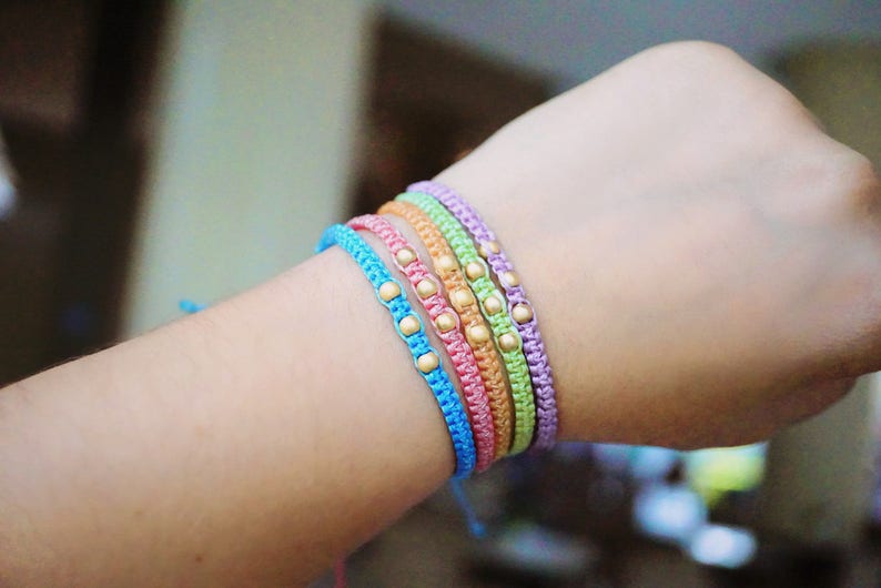 Pu&ograve; includere: Un set di cinque braccialetti dell'amicizia colorati con perline dorate. I braccialetti sono realizzati con cordini sottili e intrecciati in tonalit&agrave; di blu, verde, rosa e viola.