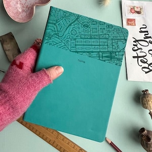 May include: Teal notebook with a map design on the top half. The word "home" is printed in the lower right corner. The notebook is held by a person wearing a pink fingerless glove. A wooden ruler and pencil are visible.