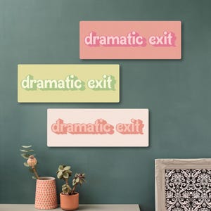 May include: Three rectangular wall art pieces with the words "dramatic exit" in a retro font. The top piece is pink, the middle is light green, and the bottom is light pink. The art is displayed on a teal wall.