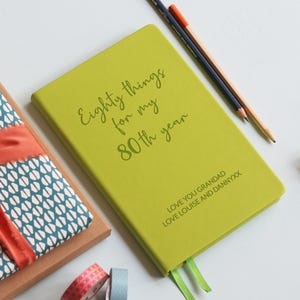 Può includere: Un taccuino verde lime con il testo "Eighty things for my 80th year" e "LOVE YOU GRANDAD LOVE LOUISE AND DANNYXX" stampato sulla copertina, accompagnato da due matite, una scatola rivestita in tessuto fantasia e due piccoli rotoli di nastro decorativo.