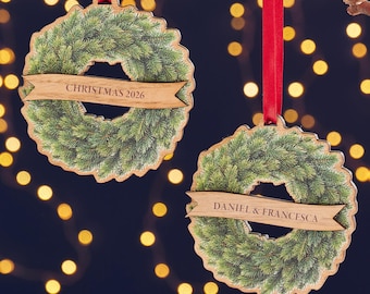 May include: Two wooden Christmas ornaments shaped like wreaths with green foliage. Each has a wooden banner with text: one says "CHRISTMAS 2026" and the other "DANIEL & FRANCESCA". Red ribbons are attached for hanging.