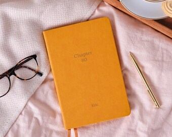 Könnte beinhalten: Ein offenes, senffarbenes Notizbuch mit dem Text "Chapter 80" und dem Namen "Eric" auf dem Einband. Ein goldener Stift liegt auf einer rosa Oberfläche neben dem Notizbuch. Eine Brille mit Schildpattmuster befindet sich links.