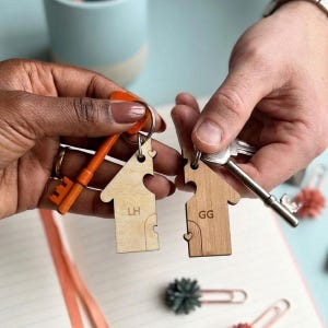 May include: Two wooden house keyrings, each with a key attached. One is light-coloured with the initials "LH", the other is darker with "GG". Both have a heart-shaped cutout and a puzzle piece design.