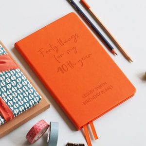 Peut inclure: Un journal orange vif avec l'inscription "Forty things for my 40th year". Il comporte deux marque-pages en ruban orange. Le texte "Lesley Smith Birthday Plans" est imprimé en dessous. Des crayons et du ruban décoratif sont également visibles.