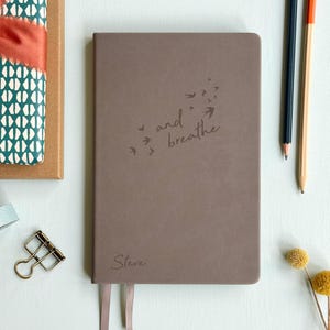 May include: A brown journal with the words "and breathe" and a flock of birds etched on the cover. The name "Steve" is at the bottom. A ribbon bookmark and two pencils are visible. Other stationery items are in the background.