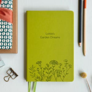 May include: A bright green notebook featuring the text "Lottie's Garden Dreams" and a floral design. The notebook has a green ribbon bookmark. Pencils, a binder clip, and a dried flower are also visible.
