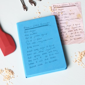 May include: A blue notebook with a handwritten recipe for "Mum's Oatie Biscuits" next to a vintage recipe card. A red spatula and silver spoons are visible, along with oats. The recipe includes measurements in ounces and teaspoons.