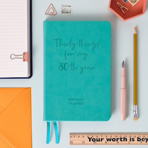 May include: A teal birthday planner with the text "Thirty things for my 30th year" and "BIRTHDAY PLANNER". A pink pen, yellow pencil, and ruler with text are also visible.