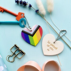 May include: Two wooden heart-shaped keychains. One keychain is rainbow colored with a chevron pattern and the other is white with the text "So proud of you xx" in gold lettering.