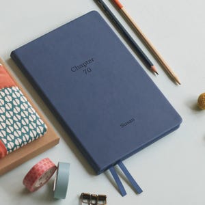May include: A navy blue journal with the text "Chapter 70" and "Susan" embossed on the cover. Two blue ribbon bookmarks extend from the top edge. Pencils, washi tape, and a small decorative item are also visible.
