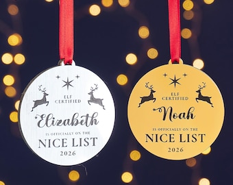 Puede incluir: Dos adornos navideños redondos con cintas rojas. Uno es plateado con el nombre "Elizabeth", el otro dorado con "Noah". Ambos tienen renos, estrellas y el texto "ELF CERTIFIED IS OFFICIALLY ON THE NICE LIST 2026."