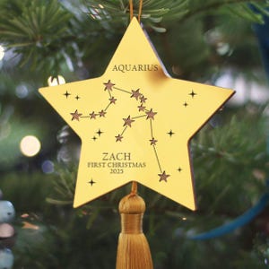 May include: A gold star-shaped Christmas ornament with the word "AQUARIUS" at the top. The ornament features a constellation design and the text "ZACH FIRST CHRISTMAS 2025". A gold tassel hangs from the bottom.