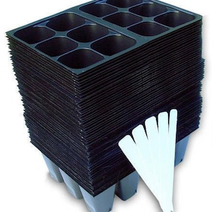 May include: A stack of black plastic seed starting trays with multiple cells, designed for planting seeds. The trays are supported by gray bases and accompanied by a bundle of white plant markers, ideal for gardening and starting seedlings.
