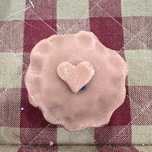 Op de afbeelding: Een lichtroze, hartvormige wax melt met een kleiner hart in het midden. De wax melt ligt op een rood en beige geruite stof.