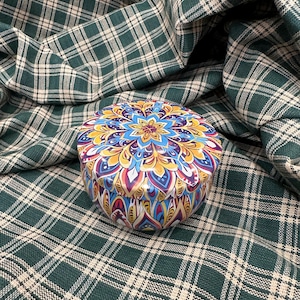 Op de afbeelding: Een ronde, kleurrijke container met een bloemenontwerp in blauw, geel en rood. De container staat op een groen-witte geruite stof. Het deksel heeft een symmetrisch patroon. Ideaal voor opslag of als decoratie.
