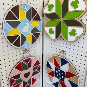 May include: Four wooden decorative signs with geometric designs. One is round with a bunny, another is a four-leaf clover, and two are oval-shaped with heart and star patterns. Each sign has a twine loop for hanging.