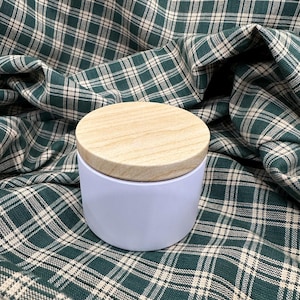 Op de afbeelding: Een kleine, ronde, witte container met een licht houten deksel. De container staat op een groen-witte geruite stof. Het deksel is gemaakt van lichtgekleurd hout.