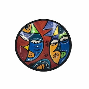 May include: A round ceramic plate with a colorful abstract design featuring three stylized faces in shades of blue, yellow, green, and red.
