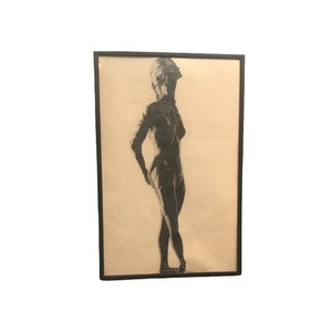 May include: A framed charcoal drawing of a person in a standing pose, looking upwards. The artwork is in grayscale, with a black frame. The figure's form is detailed, with shading that defines the body's contours. The background is a light beige.