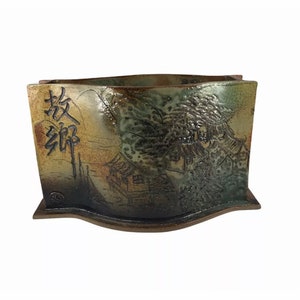 May include: A ceramic planter with a wavy, rectangular shape. The planter is decorated with a landscape scene in shades of brown, green, and white. The scene includes a mountain range, trees, and a small house. The planter has a Japanese inscription on the side.