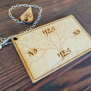 May include: A wooden Ouija board with a tree design and the words "YES" and "NO" printed on it. A silver chain with a silver pendant is attached to the board.