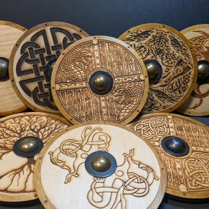 May include: A collection of seven circular wooden shields featuring intricate pyrography designs including Celtic knots, dragons, and a tree. Each shield has a central metallic boss and a light wood finish with dark burnt wood detailing.
