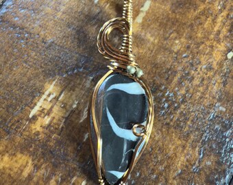 May include: A handcrafted pendant featuring a teardrop-shaped stone with black and white patterns, encased in copper wire. The pendant hangs from a black cord. The wirework has intricate details and a polished finish, showcasing the natural stone.