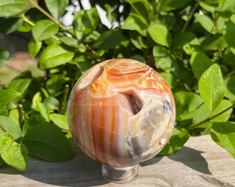 May include: A polished, spherical gemstone with bands of orange, white, and gray. The orb is displayed on a clear, round stand, set against a backdrop of green foliage. The stone's surface has natural cavities.