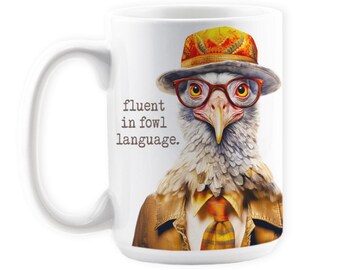 May include: White ceramic mug with a brown and white bird wearing a red, yellow, and orange hat and glasses. The mug has the text "fluent in fowl language." printed on it.