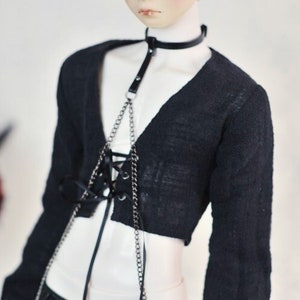 May include: A doll wearing a black cropped cardigan with long sleeves, black leather shorts, and a silver chain choker with a black leather strap. The doll has short, silver hair.