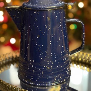 以下が含まれることがあります： 白の斑点模様が入った、ネイビーブルーのエナメルコーヒーポット。ポットには蓋、注ぎ口、ハンドルが付いています。金色の装飾トレイの上に置かれています。背景はカラフルな光でぼやけています。