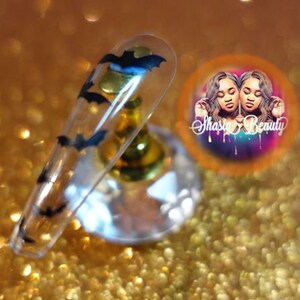 Op de afbeelding: Een heldere acrylnagel met een Halloween-thema. De nagel heeft zwarte vleermuis- en gouden maanontwerpen. De nagel wordt weergegeven op een gouden en heldere standaard, met een cirkelvormig logo op de achtergrond met de tekst "Shasia Beauty". De achtergrond is een gouden glitter.
