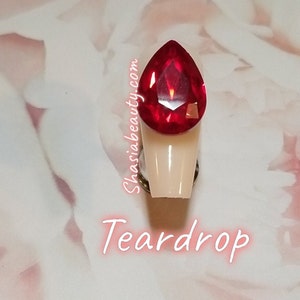 May include: A teardrop-shaped red rhinestone on a white nail tip. The nail tip is attached to a silver base. The text "Shasia Beauty.com" and "Teardrop" are visible below the nail tip.