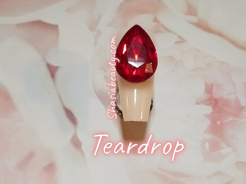 May include: A teardrop-shaped red rhinestone on a white nail tip. The nail tip is attached to a silver base. The text "Shasia Beauty.com" and "Teardrop" are visible below the nail tip.