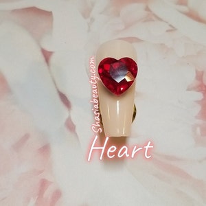 May include: A close-up of a long, light pink artificial nail with a red heart-shaped rhinestone on top. The text "ShasiaBeauty.com" and "Heart" are visible below the nail.