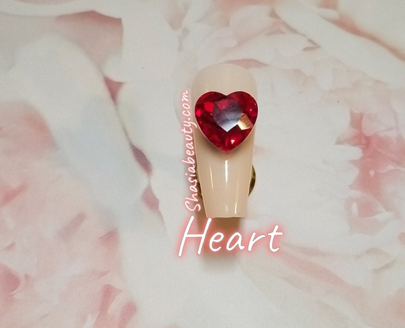 May include: A close-up of a long, light pink artificial nail with a red heart-shaped rhinestone on top. The text "ShasiaBeauty.com" and "Heart" are visible below the nail.