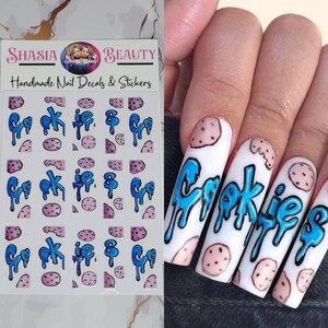 Puede incluir: Calcomanías y pegatinas para uñas con temática de galletas. La hoja de pegatinas tiene la palabra "Cookies" en fuente azul goteante, junto con ilustraciones de galletas. Las uñas están pintadas de blanco con la palabra "Cookies" en fuente azul goteante e ilustraciones de galletas.