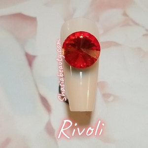 May include: A close-up of a red Rivoli rhinestone on a white nail. The nail is long and oval shaped. The rhinestone is sparkling and has a gold backing. The text "Shasia Beauty.com" is written on the nail. The text "Rivoli" is written below the nail.