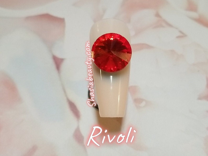 May include: A close-up of a red Rivoli rhinestone on a white nail. The nail is long and oval shaped. The rhinestone is sparkling and has a gold backing. The text "Shasia Beauty.com" is written on the nail. The text "Rivoli" is written below the nail.