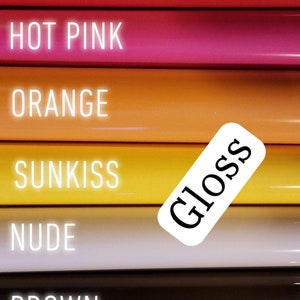 Op de afbeelding: Een close-up van een stapel glanzende vinylrollen in verschillende kleuren, waaronder rood, felroze, oranje, geel, nude, bruin en ultraviolet. Elke rol heeft een label met de kleurnaam. Een wit label met het woord "Gloss" is ook zichtbaar.