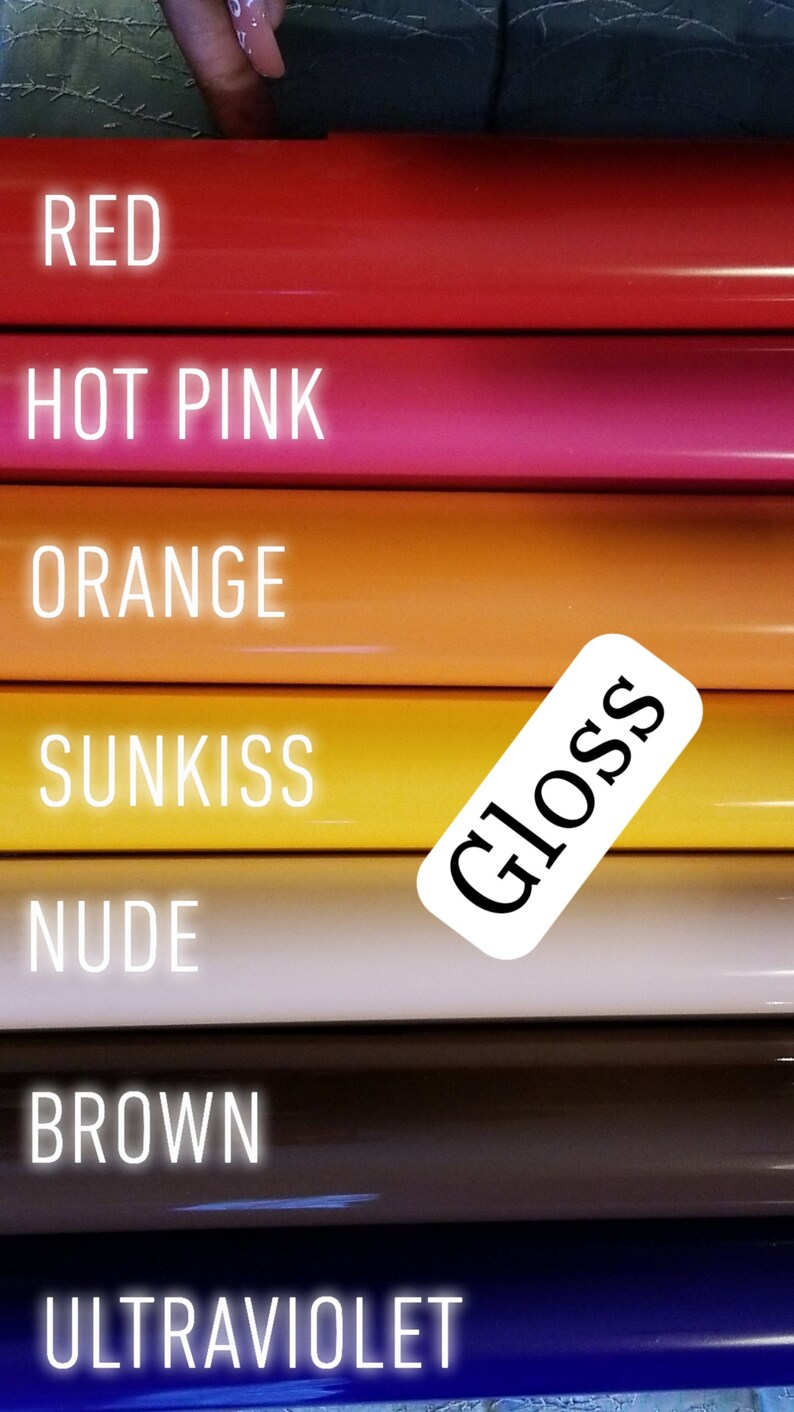 Op de afbeelding: Een close-up van een stapel glanzende vinylrollen in verschillende kleuren, waaronder rood, felroze, oranje, geel, nude, bruin en ultraviolet. Elke rol heeft een label met de kleurnaam. Een wit label met het woord "Gloss" is ook zichtbaar.