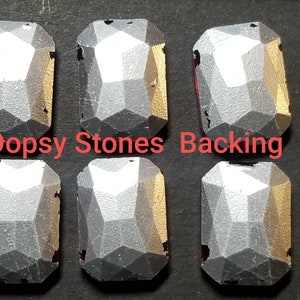 May include: Six silver-colored, faceted, rectangular-shaped stones with a flat back. The stones are arranged in two rows of three. The text "Oopsy Stones Backing" is printed in red below the stones.