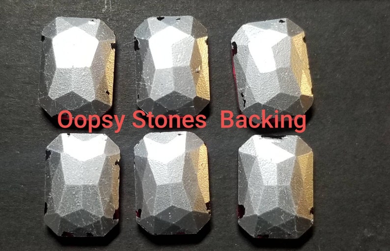 May include: Six silver-colored, faceted, rectangular-shaped stones with a flat back. The stones are arranged in two rows of three. The text "Oopsy Stones Backing" is printed in red below the stones.