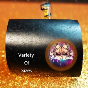 Op de afbeelding: Zwart, gebogen product met een vleermuismotief en de woorden "Variety Of Sizes". Een cirkelvormige afbeelding met twee vrouwen en de tekst "Shasia Beauty" staat op het product. De achtergrond is oranje en glinsterend.