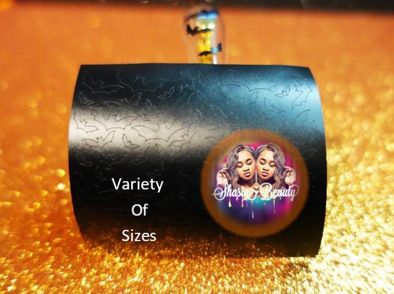 Op de afbeelding: Zwart, gebogen product met een vleermuismotief en de woorden "Variety Of Sizes". Een cirkelvormige afbeelding met twee vrouwen en de tekst "Shasia Beauty" staat op het product. De achtergrond is oranje en glinsterend.