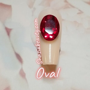 May include: A close-up of a single oval-shaped fake nail with a large red rhinestone on top. The nail is a light beige color. The text "Shasiabeauty.com" is written vertically on the side of the nail. The text "Oval" is written below the nail.