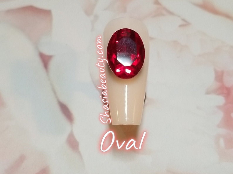 May include: A close-up of a single oval-shaped fake nail with a large red rhinestone on top. The nail is a light beige color. The text "Shasiabeauty.com" is written vertically on the side of the nail. The text "Oval" is written below the nail.