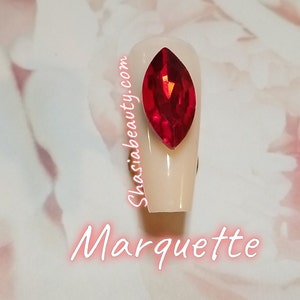 May include: A close-up of a white nail with a large, red, faceted, marquise-shaped rhinestone. The nail is surrounded by a white background with the text "Shasiabeauty.com" and "Marquette" written in pink.